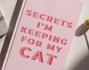Op de afbeelding: Een lichtroze notitieboek met harde kaft en de tekst "SECRETS I'M KEEPING FOR MY CAT" in dikke, gelaagde rode en roze hoofdletters. Het boek heeft een matte afwerking en is schuin geplaatst, waardoor de voorkant van de kaft volledig zichtbaar is.