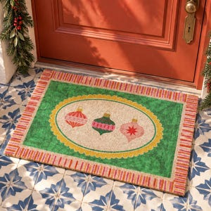 May include: A rectangular doormat featuring a green center with a beige oval frame containing three Christmas ornaments in red, green, and pink. The mat has a striped red and white border and is placed on a tiled floor in front of a red door.