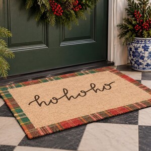 Puede incluir: Un felpudo rectangular con un centro de color tostado natural que presenta el texto negro en cursiva hohoho. La alfombrilla est&aacute; enmarcada por un borde de tela de cuadros rojos y verdes. El dise&ntilde;o es sencillo y funcional para la entrada.