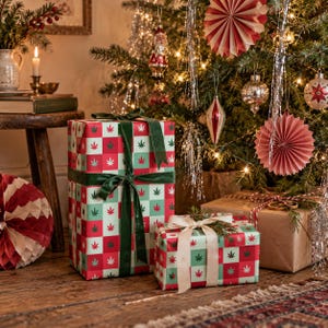 May include: Two rectangular gift boxes wrapped in red and light green checkered paper featuring a repeating cannabis leaf pattern. The taller box is tied with a dark green velvet ribbon, while the smaller box has a cream-coloured satin ribbon.