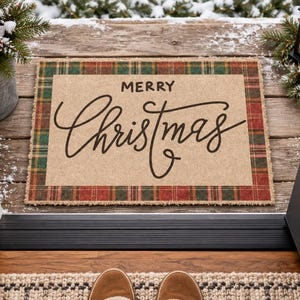 May include: A rectangular doormat with a tan center featuring the black text Merry Christmas in a script font. The mat has a red and green plaid border with a frayed edge. The design is centered on a wooden porch with a snowy outdoor setting.