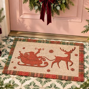 K&ouml;nnte beinhalten: Eine rechteckige Fu&szlig;matte mit naturfarbener Mitte und einem roten Motiv von einem Weihnachtsmann im Schlitten, der von einem Rentier gezogen wird. Die Matte hat einen rot-gr&uuml;nen Karorand und ist mit kleinen roten Schneeflocken verziert.