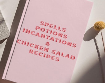 Op de afbeelding: Een lichtroze hardcover boek met de tekst SPELLS POTIONS INCANTATIONS & CHICKEN SALAD RECIPES gedrukt in een vet, gelaagd rood en roze lettertype op de voorkant. Het boek ligt in een hoek met een kleine gele bloem ernaast.