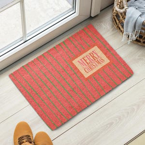 May include: A rectangular doormat with a textured red surface featuring thin diagonal green stripes. A central tan rectangular patch displays the text MERRY CHRISTMAS in a red, stylized font. The mat has a coarse, fibrous appearance.