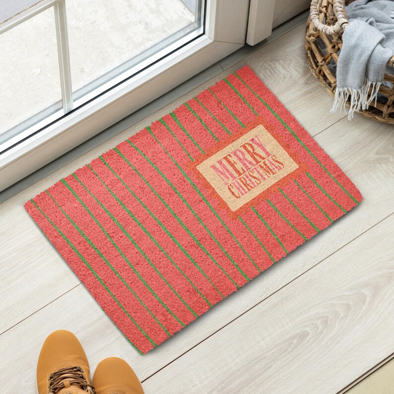 May include: A rectangular doormat with a textured red surface featuring thin diagonal green stripes. A central tan rectangular patch displays the text MERRY CHRISTMAS in a red, stylized font. The mat has a coarse, fibrous appearance.