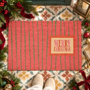 May include: A rectangular coir doormat with a textured red surface featuring thin vertical green stripes. A central rectangular beige label with a decorative orange border displays the text Merry Christmas in red and pink lettering.