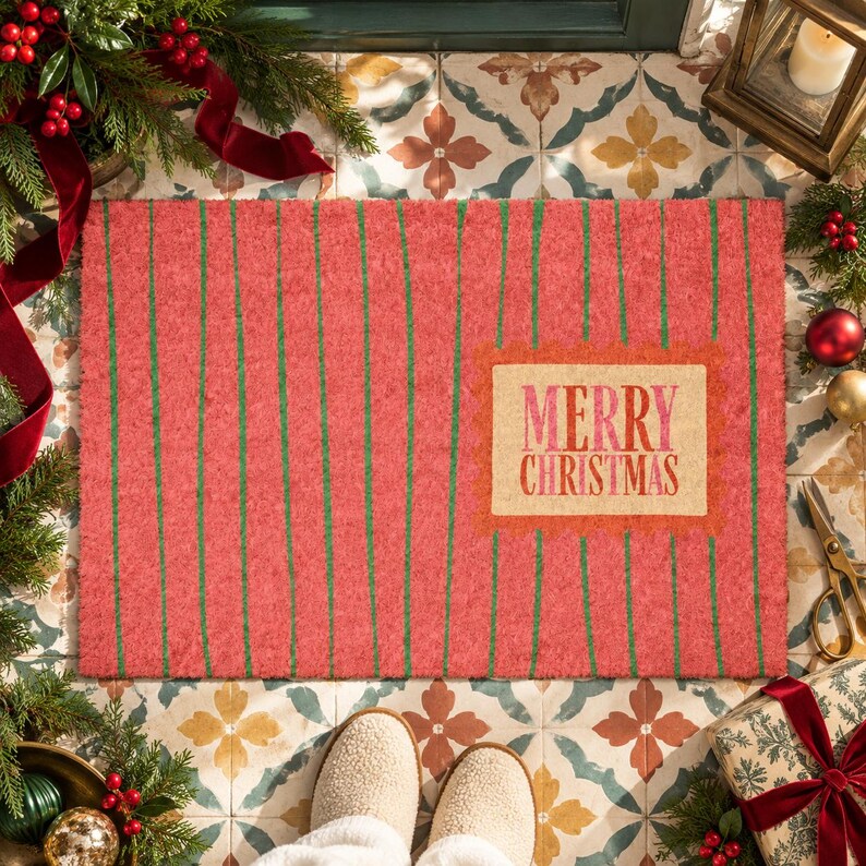May include: A rectangular coir doormat with a textured red surface featuring thin vertical green stripes. A central rectangular beige label with a decorative orange border displays the text Merry Christmas in red and pink lettering.