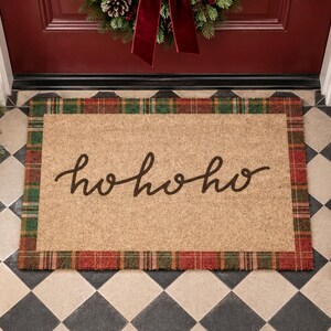 Puede incluir: Un felpudo rectangular de fibra de coco con un centro de color tostado natural y el texto hohoho escrito en una caligraf&iacute;a cursiva de color marr&oacute;n oscuro. La alfombra est&aacute; enmarcada por un borde decorativo con un patr&oacute;n de cuadros rojo y verde.