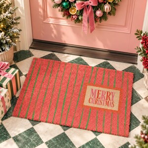 May include: A rectangular coir doormat featuring a vibrant red base with thin vertical green stripes. A central rectangular tan inset displays the text Merry Christmas in bold red and pink lettering, creating a festive holiday entrance accent.
