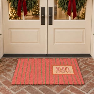 May include: A rectangular doormat featuring a red base with thin vertical green stripes. A rectangular tan patch is positioned on the right side of the mat with the text Merry Christmas printed in a red font across the center of the patch.