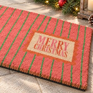 May include: A rectangular coir doormat featuring a red background with vertical green stripes. In the center, a tan rectangular patch with a scalloped orange border displays the text MERRY CHRISTMAS in bold, dark pink capital letters.