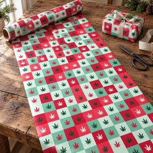 May include: A roll of wrapping paper featuring a repeating checkerboard pattern of red and light green squares, each printed with a small cannabis leaf icon in a contrasting colour. A gift box wrapped in the same paper sits nearby with scissors.