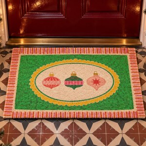 May include: A rectangular doormat featuring a green border with a pink and white striped edge. The center is a cream-colored oval with a yellow scalloped frame, displaying three hanging Christmas ornaments in red, green, and pink colors.