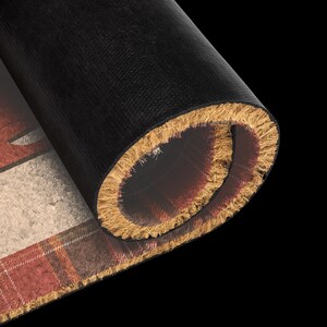 Puede incluir: Un felpudo rectangular con un borde enrollado, que presenta un patr&oacute;n de cuadros rojos y marrones en la superficie. La parte inferior de la alfombra es negra y lisa, mientras que la parte superior tiene una textura fibrosa y &aacute;spera.