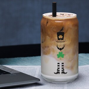 Könnte beinhalten: Ein klares, dosenförmiges Glas, gefüllt mit einem geschichteten Eiskaffeegetränk. Der Becher zeigt eine schwarze Grafik eines Leprechauns mit Hut, Bart und Kleeblatt. Ein schwarzer Strohhalm ist in das Getränk gesteckt. Der Becher steht auf einer weißen Oberfläche.