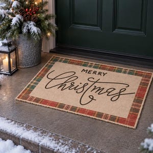 May include: A rectangular doormat with a tan center featuring the black text Merry Christmas in a script font. The mat is bordered by a red, green, and tan plaid pattern. The doormat is positioned on a concrete porch in front of a dark green door.
