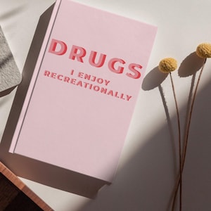 Op de afbeelding: Een roze notitieboek met het woord "DRUGS" in rode, 3D-achtige letters en de zin "I ENJOY RECREATIONALLY" in kleinere rode tekst. Het notitieboek ligt op een wit oppervlak met gedroogde gele bloemen.