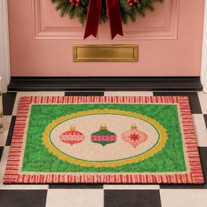 May include: A rectangular doormat featuring a central cream-colored oval with three decorative Christmas ornaments in red, green, and pink. The mat has a bright green border surrounded by a red and pink striped edge, creating a festive look.