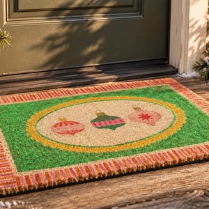 May include: A rectangular coir doormat featuring a green border with a red and orange striped edge. The center has a tan oval framed by a yellow scalloped border, displaying three decorative Christmas ornaments in red, green, and pink hues.