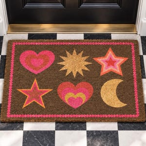 May include: A rectangular brown coir doormat featuring a bright pink border and a pattern of six celestial and heart-shaped icons. The design includes a pink heart, a gold sun, a multi-colored star, a second star, a layered heart, and a gold crescent moon.