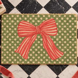 May include: A rectangular doormat featuring a green background with a repeating pattern of small cream-coloured polka dots. Centered on the mat is a large, prominent red bow with thin, vertical cream-coloured stripes throughout the fabric design.