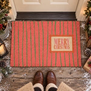 May include: A rectangular doormat with a textured coral red surface featuring vertical green stripes. A central rectangular tan patch with a scalloped orange border displays the text Merry Christmas in pink and red lettering.