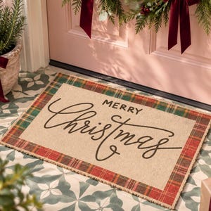 May include: A rectangular doormat with a tan center and a red and green plaid border. The text "MERRY" is printed in a simple sans-serif font above the word "Christmas" written in a large, black, flowing cursive script across the center.