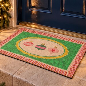 May include: A rectangular doormat featuring a green border with a red and white striped edge. The center has an oval cream-colored field with a yellow scalloped border, displaying three illustrated Christmas ornaments in pink, green, and red.