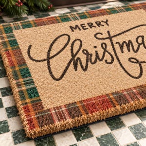 May include: A rectangular coir doormat with a natural tan center featuring the black text Merry Christmas in a script font. The border consists of a woven plaid pattern in shades of green, red, brown, and yellow, creating a textured frame.