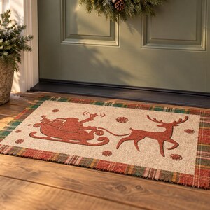 K&ouml;nnte beinhalten: Eine rechteckige Fu&szlig;matte mit beigefarbener Mitte, die eine rote Silhouette des Weihnachtsmanns im Schlitten, gezogen von einem Rentier, zeigt. Die Matte hat einen rot-gr&uuml;n-goldenen Karorand. Kleine rote Schneeflocken sind verteilt.