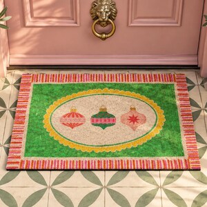 May include: A rectangular doormat featuring a bright green border with a pink and orange striped edge. The center has a beige oval with a yellow scalloped frame, displaying three hanging Christmas ornaments in red, green, and pink colors.