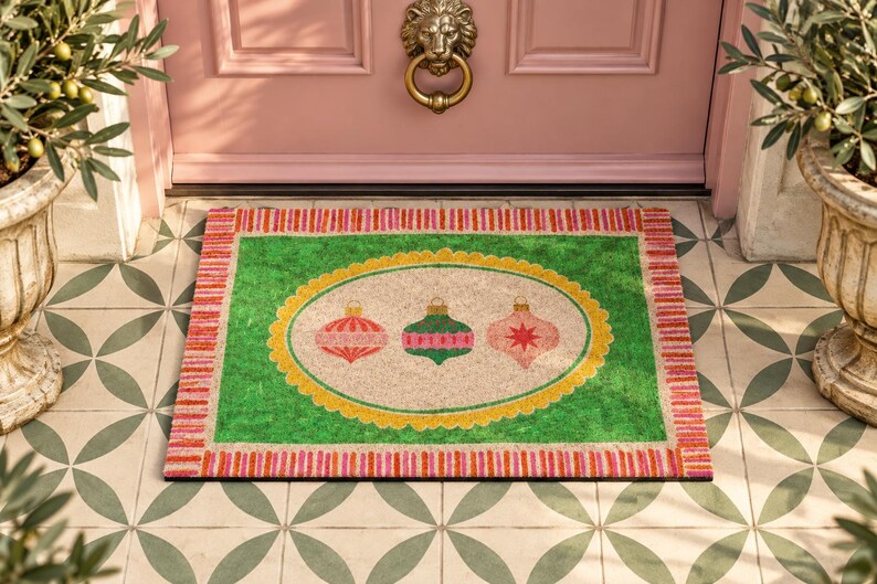 May include: A rectangular doormat featuring a bright green border with a pink and orange striped edge. The center has a beige oval with a yellow scalloped frame, displaying three hanging Christmas ornaments in red, green, and pink colors.