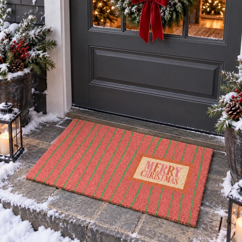 May include: A rectangular doormat featuring a red and green striped pattern with a central tan rectangular patch containing the text Merry Christmas in red lettering. The mat is positioned on a stone porch in front of a dark gray door.