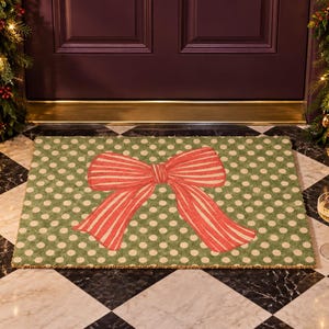 May include: A rectangular doormat featuring a green background with a pattern of small white polka dots and a large, centred red and white striped bow graphic. The mat is made of a coarse, natural fibre material with a flat, low-profile design.