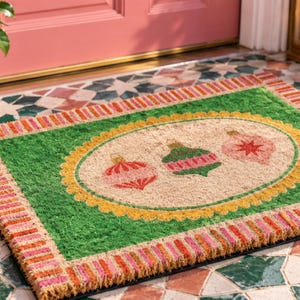 May include: A rectangular coir doormat featuring a green border with a striped pink and orange edge. The center has a beige oval with a yellow scalloped frame, displaying three illustrated Christmas ornaments in shades of pink and green.