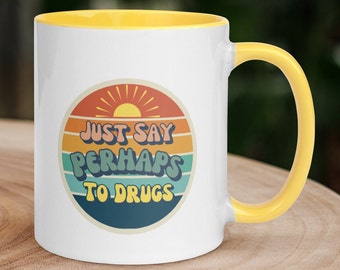 Op de afbeelding: Een witte keramische mok met een gele binnenkant en handvat. De voorzijde heeft een cirkelvormige afbeelding met een zonsondergangontwerp in oranje, gele en blauwgroene strepen. De tekst "JUST SAY PERHAPS TO DRUGS" is gedrukt in een donkerblauw lettertype.