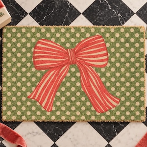 May include: A rectangular doormat featuring a green background with a repeating pattern of small cream-coloured polka dots. Centered on the mat is a large, prominent red bow with thin, vertical cream-coloured stripes throughout the fabric design.