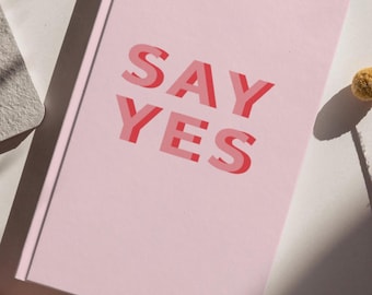 Op de afbeelding: Een lichtroze notitieboek met harde kaft en de tekst SAY YES in een vetgedrukt, gelaagd rood en roze lettertype. De kaft heeft een gladde, matte afwerking en een strak, rechthoekig ontwerp met een zichtbare rug aan de linkerkant van het boek.