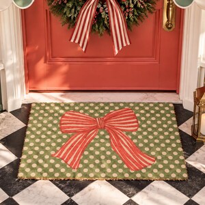May include: A rectangular doormat featuring a green background with a pattern of small white polka dots and a large, centred red and white striped bow. The mat is positioned on a black and white chequered floor in front of a red door.