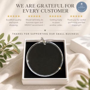 May include: A circular clear glass ornament with a faceted edge, featuring a silver-toned metal bail at the top. The ornament is presented inside a square white box with a black interior lining, displayed against a soft fabric background.