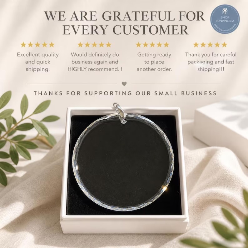 May include: A circular clear glass ornament with a faceted edge, featuring a silver-toned metal bail at the top. The ornament is presented inside a square white box with a black interior lining, displayed against a soft fabric background.
