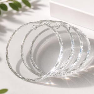 May include: A set of four clear, circular acrylic ornaments featuring a faceted, diamond-cut edge design. Each transparent disc includes a small, pre-drilled hole at the top for hanging, creating a reflective surface that catches light.