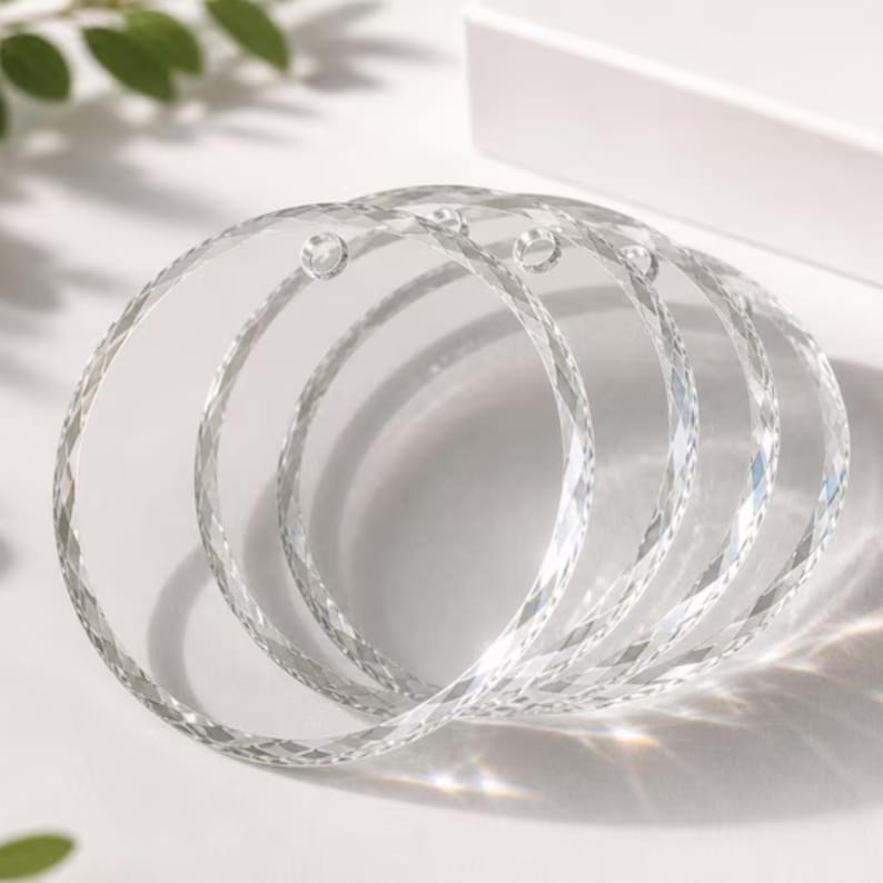 May include: A set of four clear, circular acrylic ornaments featuring a faceted, diamond-cut edge design. Each transparent disc includes a small, pre-drilled hole at the top for hanging, creating a reflective surface that catches light.