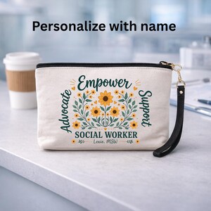 May include: Cream-colored zippered pouch with the words "Empower Advocate Support" and "Social Worker" printed on it. The design includes yellow sunflowers and green leaves. A black wrist strap is attached to the zipper.