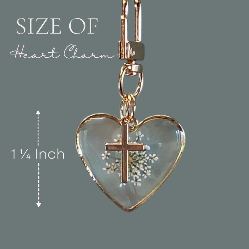 May include: A heart-shaped charm with a gold-tone metal frame, containing a white pressed flower and a small gold-tone cross. The charm hangs from a gold-tone lobster clasp. Text indicates the size of the heart charm is 1 1/4 inches.