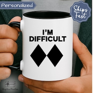 May include: White ceramic mug with a black interior and handle. The mug features the text "I'M DIFFICULT" above two black diamond shapes. The mug is held by a person.