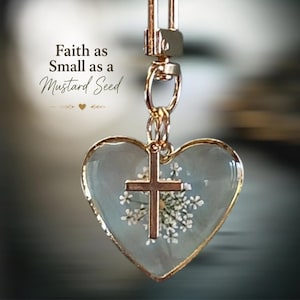 May include: A heart-shaped pendant with a gold-toned metal rim, containing a pressed white flower and a small gold-toned cross charm. The text Faith as Small as a Mustard Seed is printed above the pendant, which hangs from a gold-toned lobster clasp.