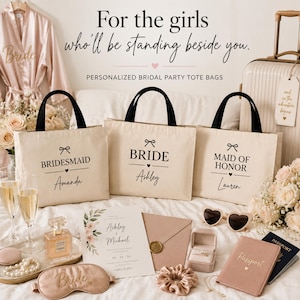 May include: Three beige canvas tote bags with black handles, each printed with a bow icon, a title, and a name. The bags read BRIDESMAID Amanda, BRIDE Ashley, and MAID OF HONOR Lauren. The scene includes a wedding invitation, perfume, and accessories.
