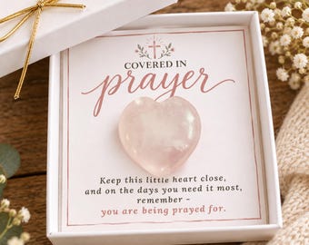 Op de afbeelding: Een gepolijste lichtroze rozenkwarts hartsteen op een witte kaart in een witte geschenkdoos. De kaart bevat een kruisicoon en de tekst: COVERED IN PRAYER. Keep this little heart close, and on the days you need it most, remember - you are being prayed for.