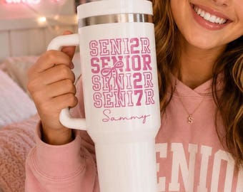 May include: A white insulated tumbler with a handle and a straw, featuring the pink text SENIOR 27 repeated in a block font with a small bow graphic and the name Sammy written in a cursive script below the text on the front of the cup.